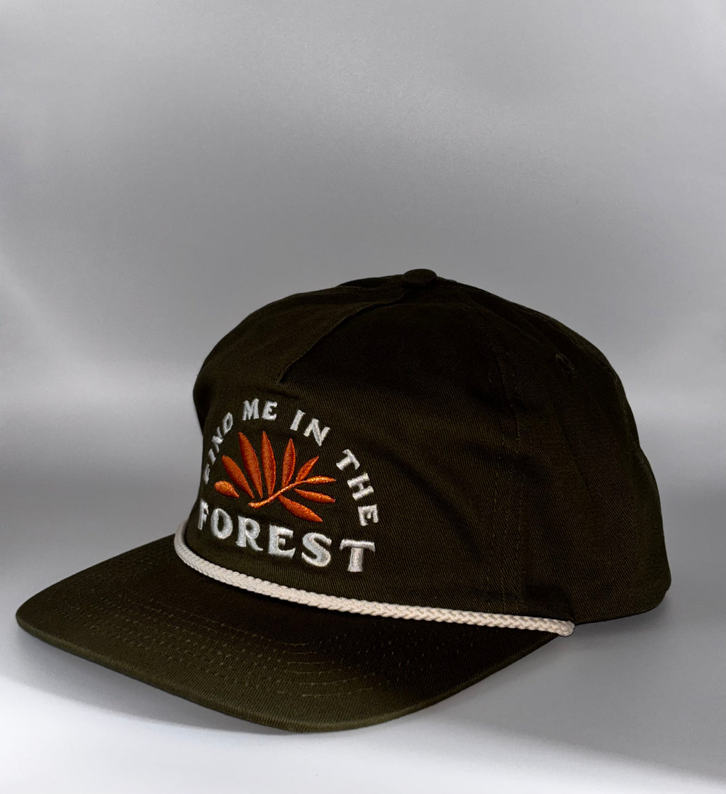 A dark olive green baseball cap with white embroidered text reading 'FIND ME IN THE FOREST' and an orange leaf design on the front, plus a white rope detail along the edge of the brim, shown against a gray studio background.