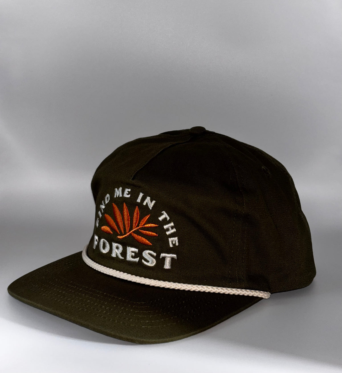 A dark olive green baseball cap with white embroidered text reading 'FIND ME IN THE FOREST' and an orange leaf design on the front, plus a white rope detail along the edge of the brim, shown against a gray studio background.