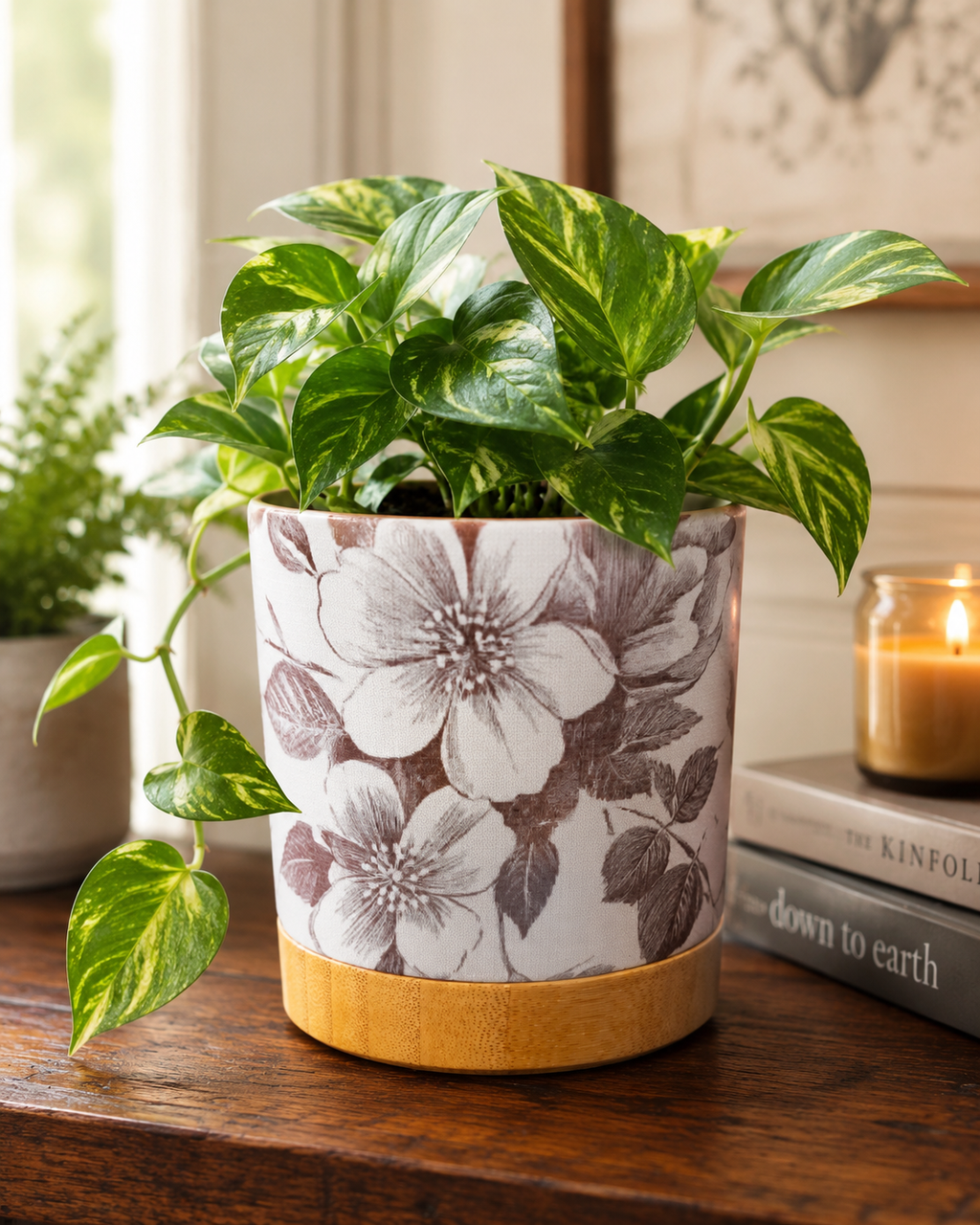 White ceramic floral planter with grey botanical print and bamboo tray, holding a trailing pothos on a wood table styled with books and a candle