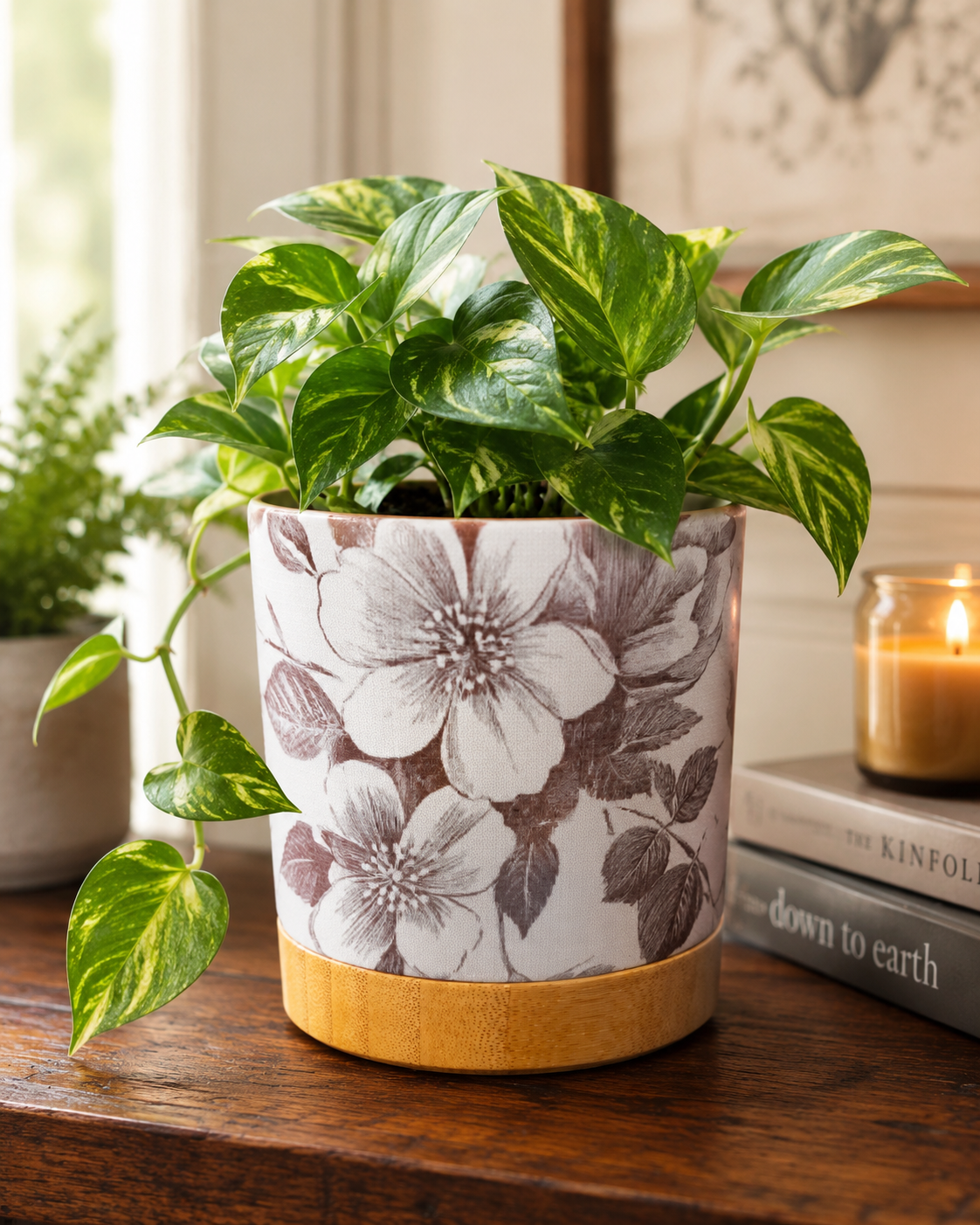 White ceramic floral planter with grey botanical print and bamboo tray, holding a trailing pothos on a wood table styled with books and a candle