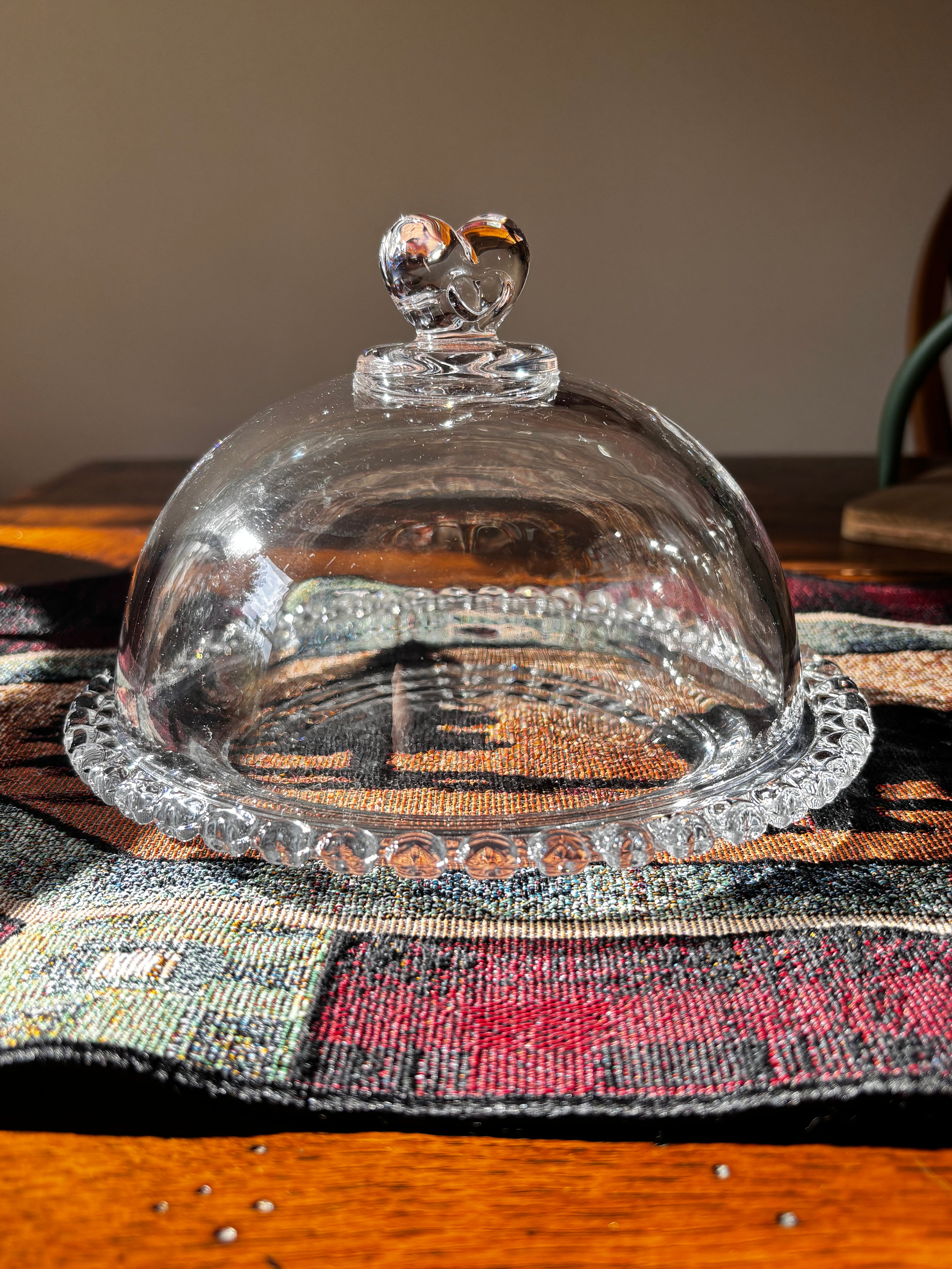 A clear glass cloche dome with a heart-shaped finial on top and a scalloped base, displayed on a patterned woven textile over a wooden surface.