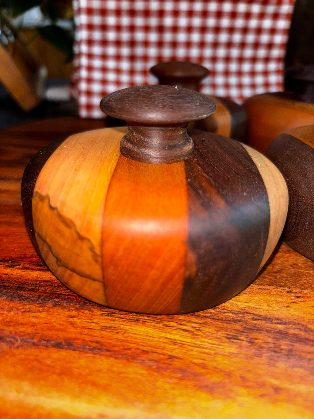 Handmade extra-large cathead biscuit cutter with a bold segmented wood body in alternating maple, cherry, and dark walnut tones, topped with a dark rounded knob, resting on a warm wood surface at a rustic artisan market."