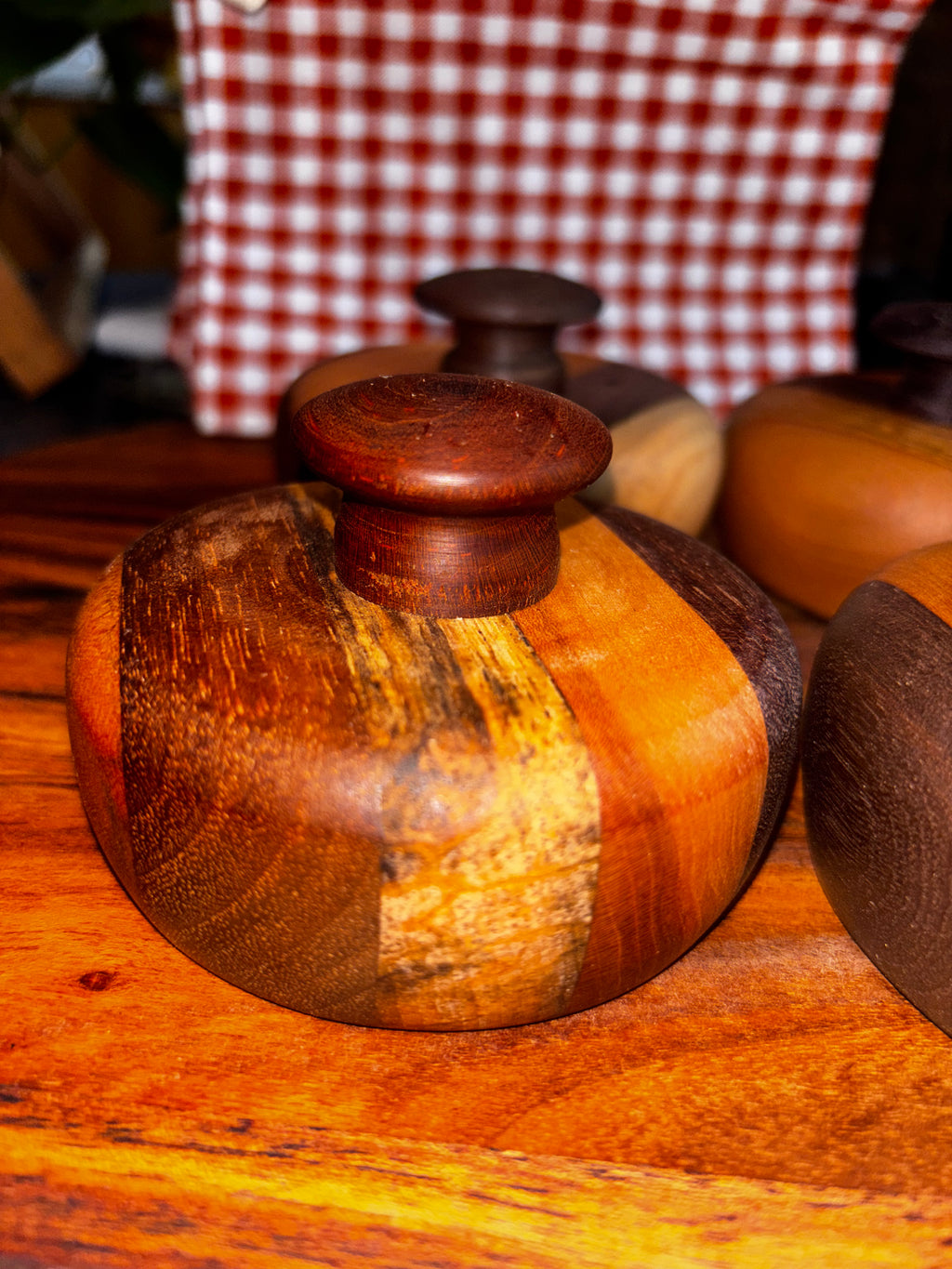 Handmade extra-large cathead biscuit cutter with a striking mixed-wood body featuring warm amber, walnut, and spalted wood grain patterns, topped with a smooth rounded cherry knob, displayed at a rustic market with red gingham in the background."