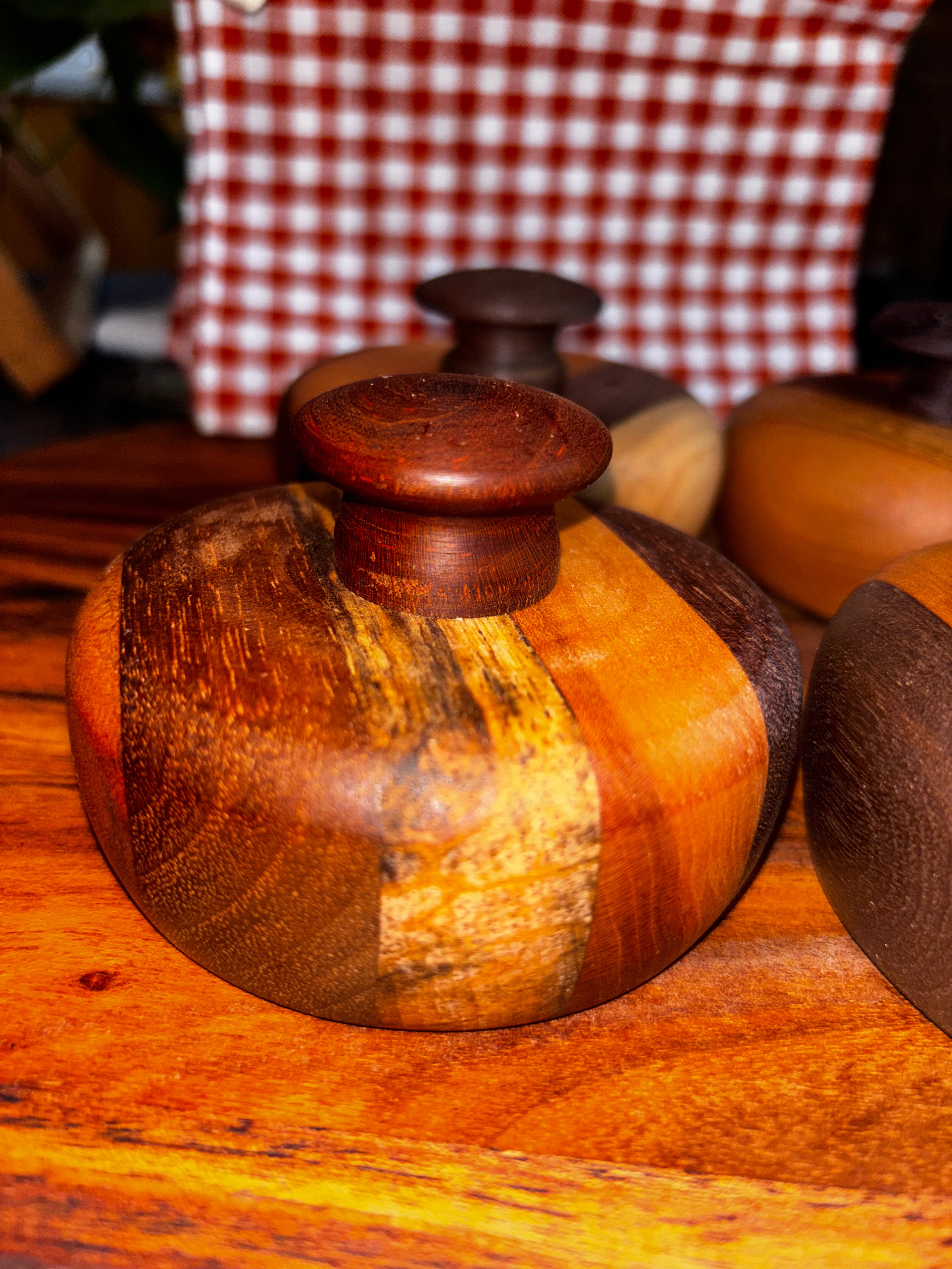 Handmade extra-large cathead biscuit cutter with a striking mixed-wood body featuring warm amber, walnut, and spalted wood grain patterns, topped with a smooth rounded cherry knob, displayed at a rustic market with red gingham in the background."