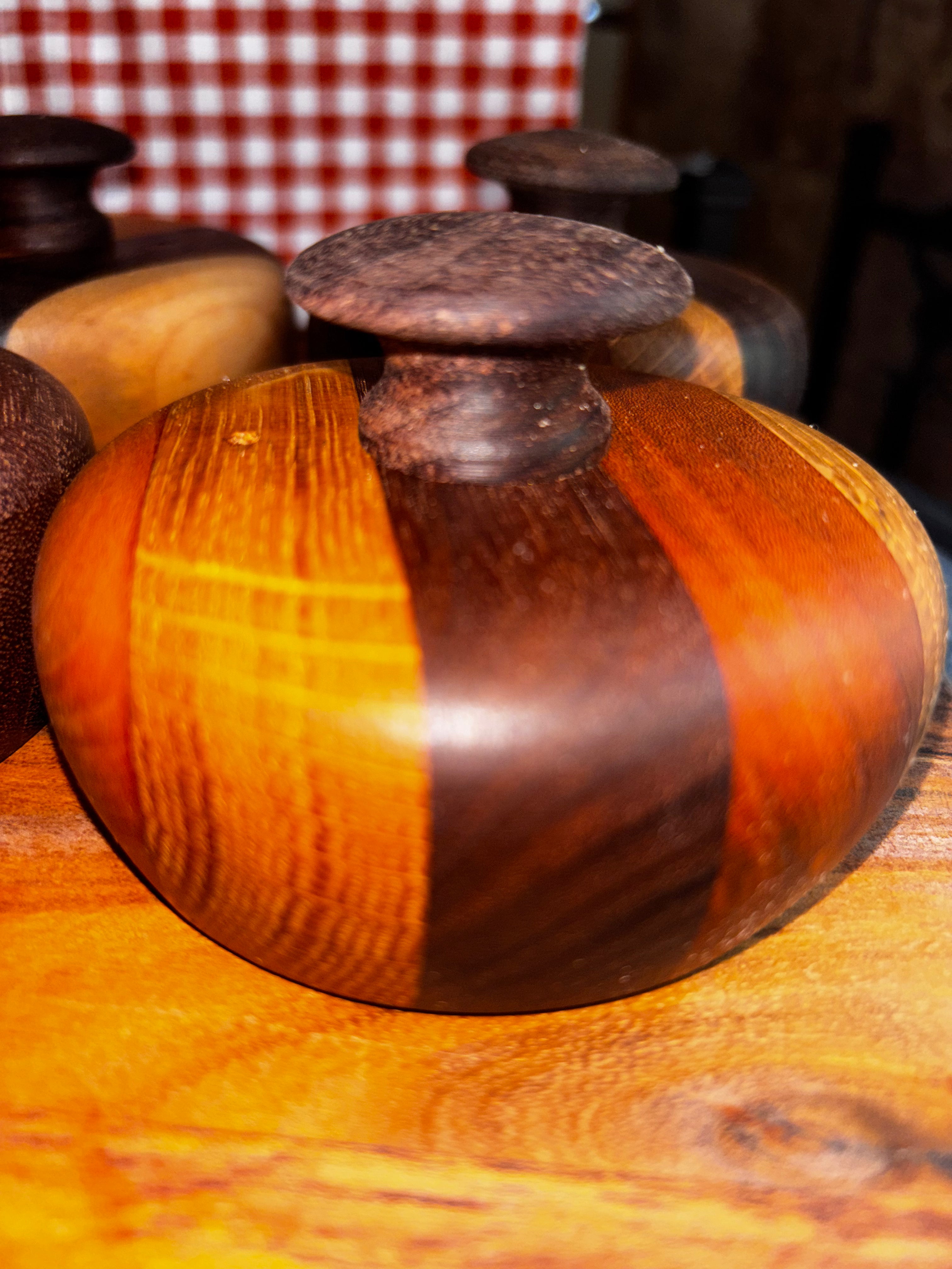 Close-up of a handmade extra-large cathead biscuit cutter resting on a wood surface, showcasing rich striped walnut and cherry wood tones with a smooth rounded knob, surrounded by additional handcrafted biscuit cutters in a rustic market setting."