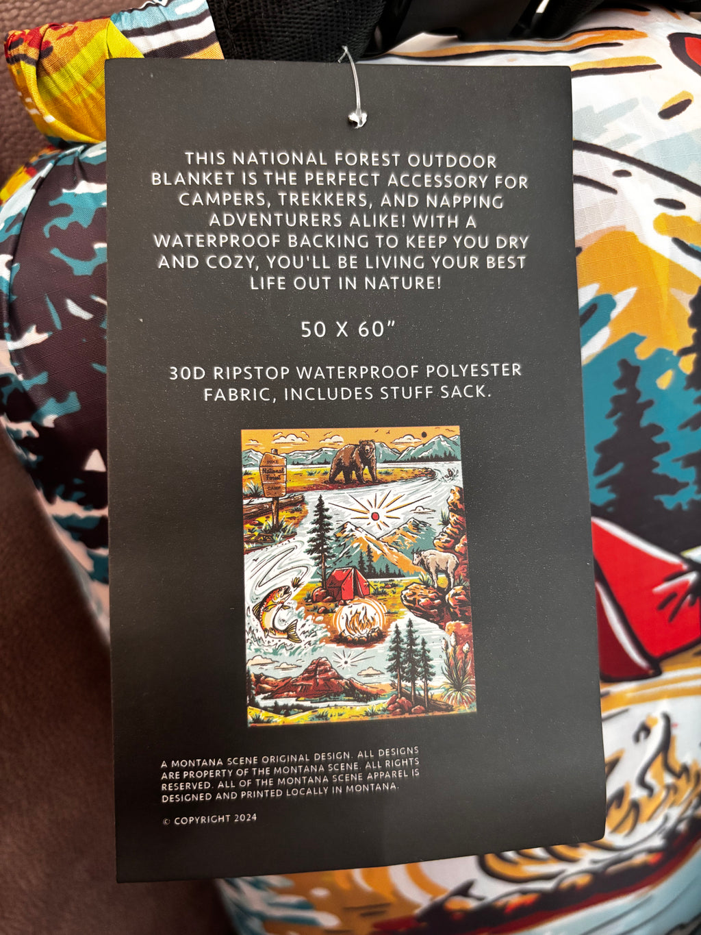 Back hang tag for The Montana Scene National Forest Outdoor Picnic Blanket — lists dimensions (50 x 60 in), 30D ripstop waterproof polyester fabric, includes stuff sack, designed and printed in Montana"