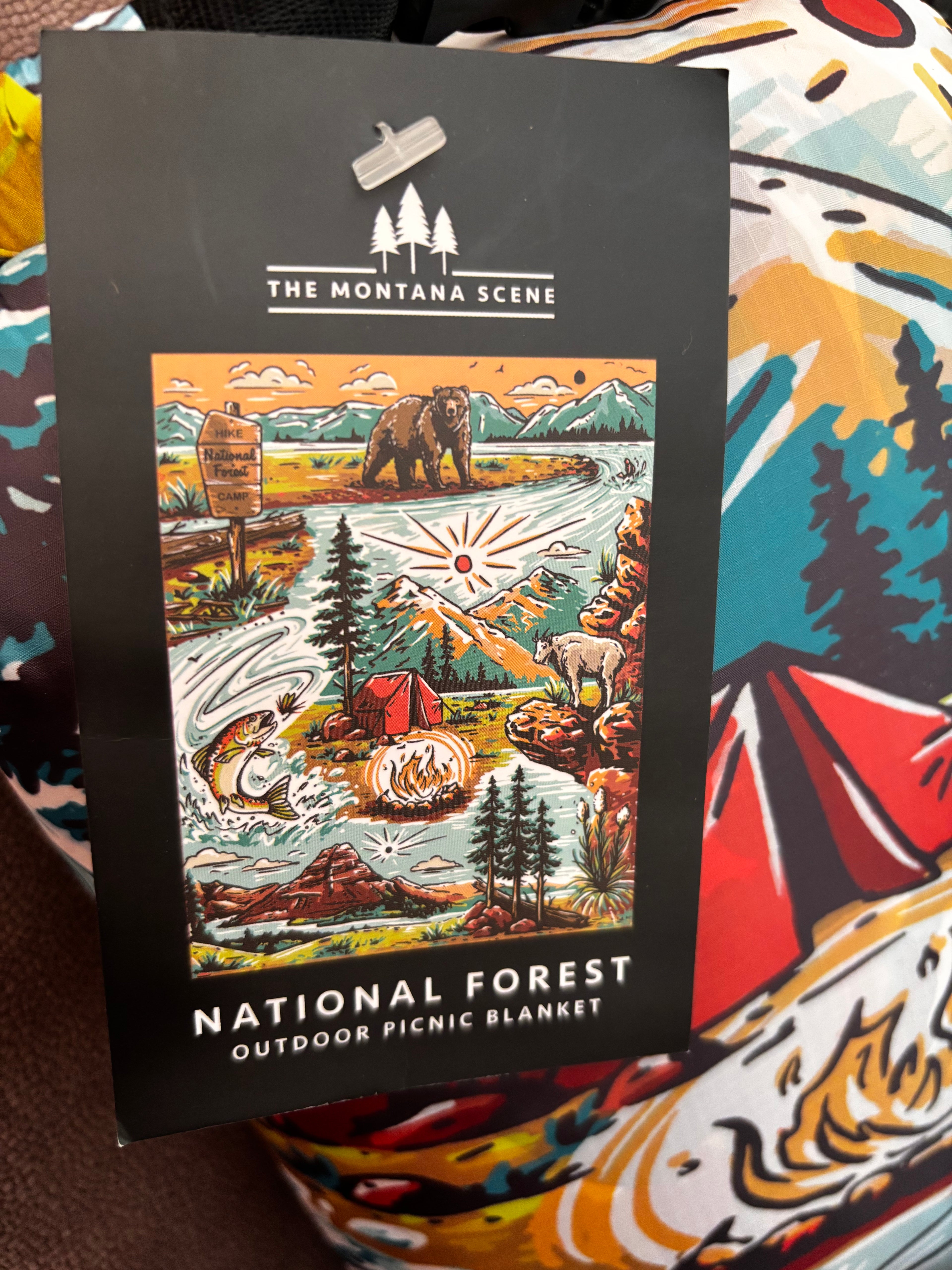 Front hang tag for The Montana Scene National Forest Outdoor Picnic Blanket — black card with illustrated camping scene featuring a bear, trout, campfire, and tent"