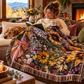 The Montana Scene Flower Meadow knit blanket draped over a woman reading on a cozy sofa by a fireplace, with a golden retriever beside her and mountain views through the window — fall lifestyle setting"