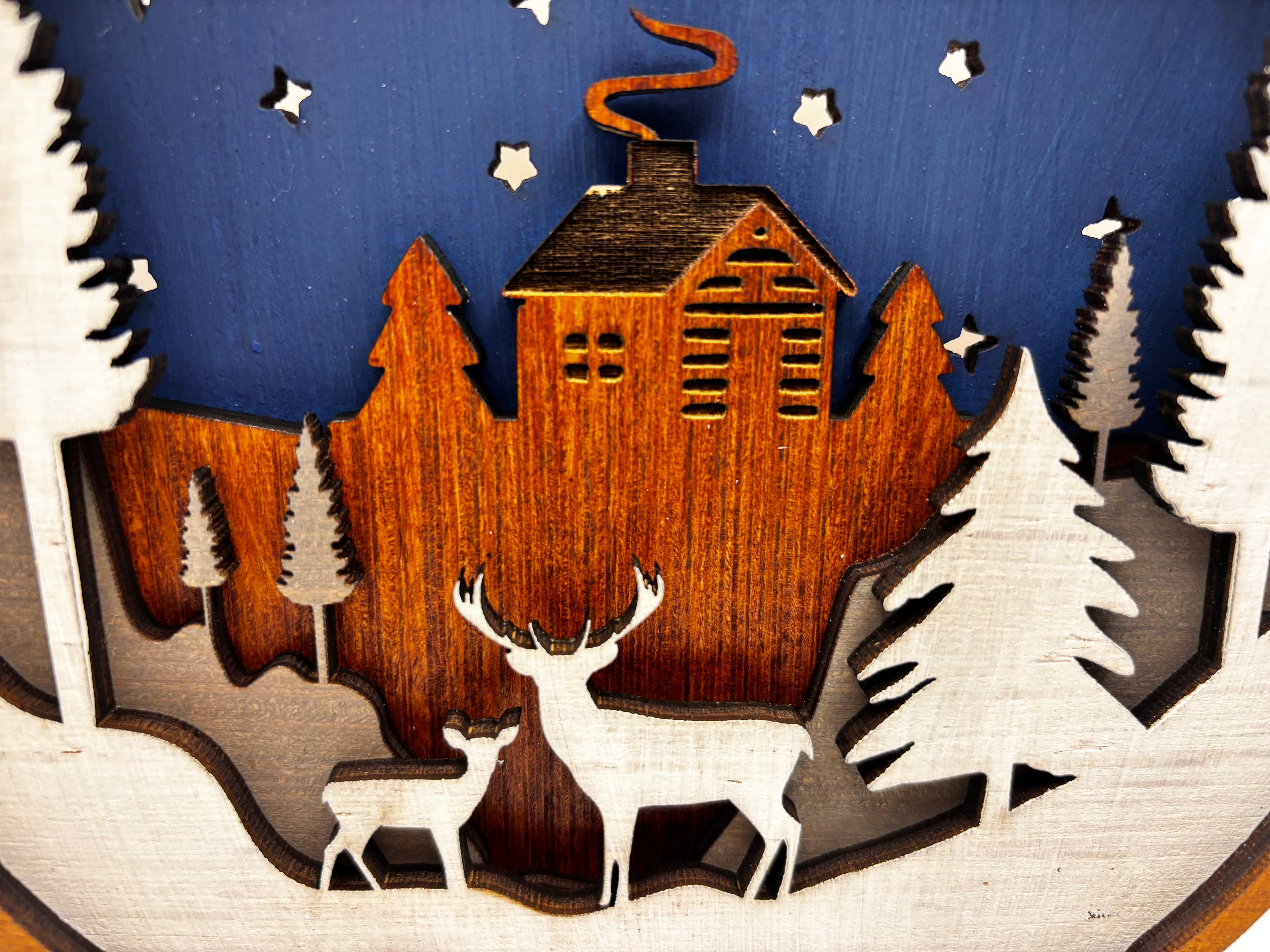 Detail of two white deer beneath a wooden cabin, surrounded by layered pine trees and snow.