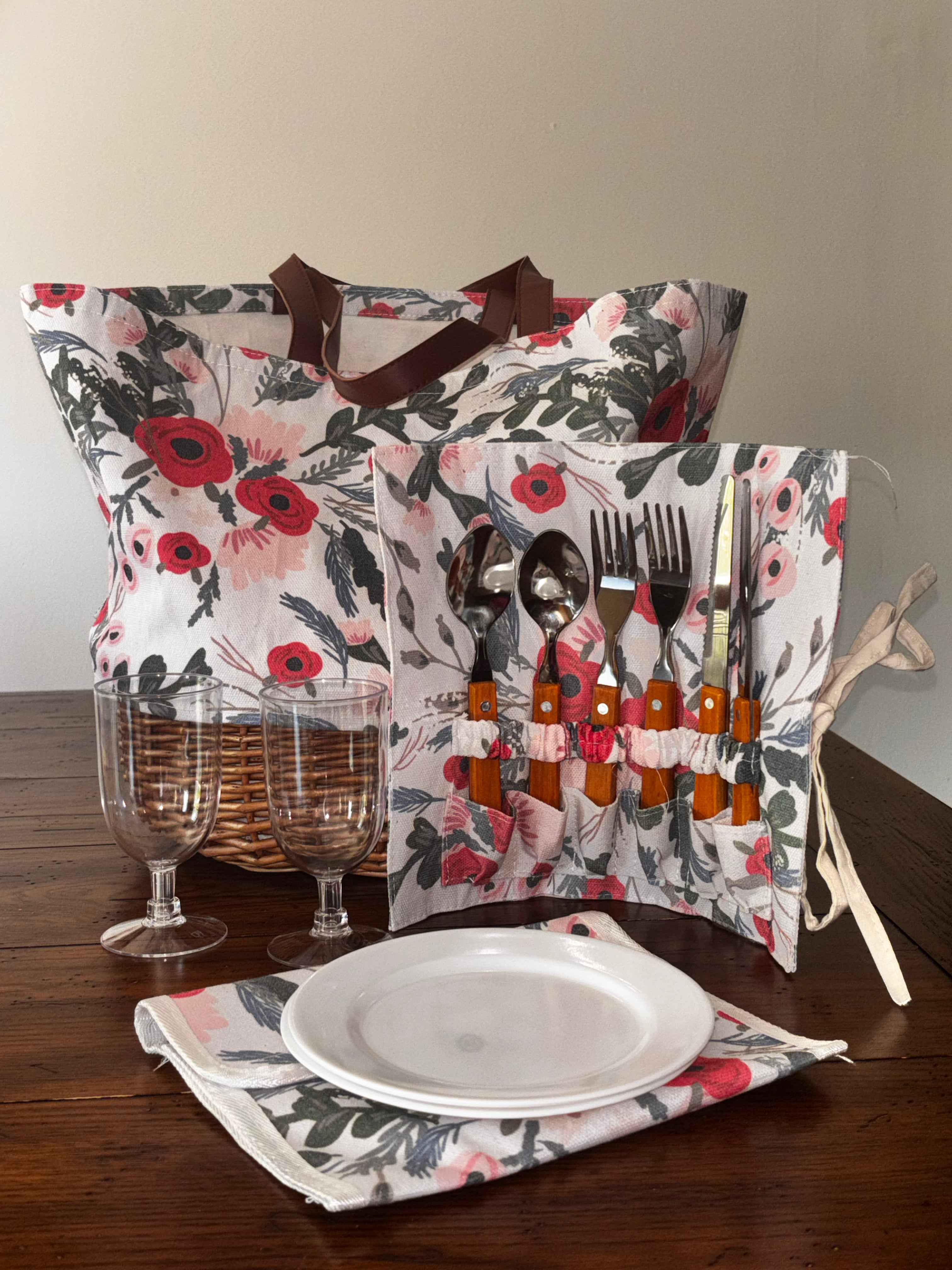 Floral Picnic Time Promenade tote with woven willow base, open cutlery holder, two clear drinking glasses, and stacked white plates.
