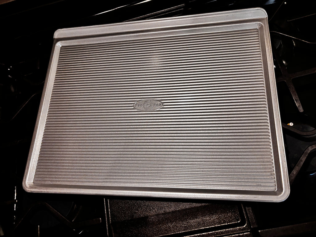 Angled view of the USA Pan large cookie sheet showing its corrugated surface, low sides and raised gripping edge.