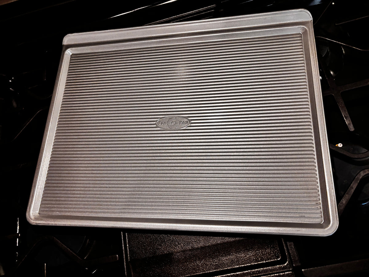 Angled view of the USA Pan large cookie sheet showing its corrugated surface, low sides and raised gripping edge.