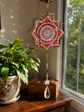  Handcrafted mandala sun catcher hanging in a sunny window, casting colorful rainbow reflections on the wall, with a green houseplant and wooden box in the background — by Sleeping Whiskers Studio