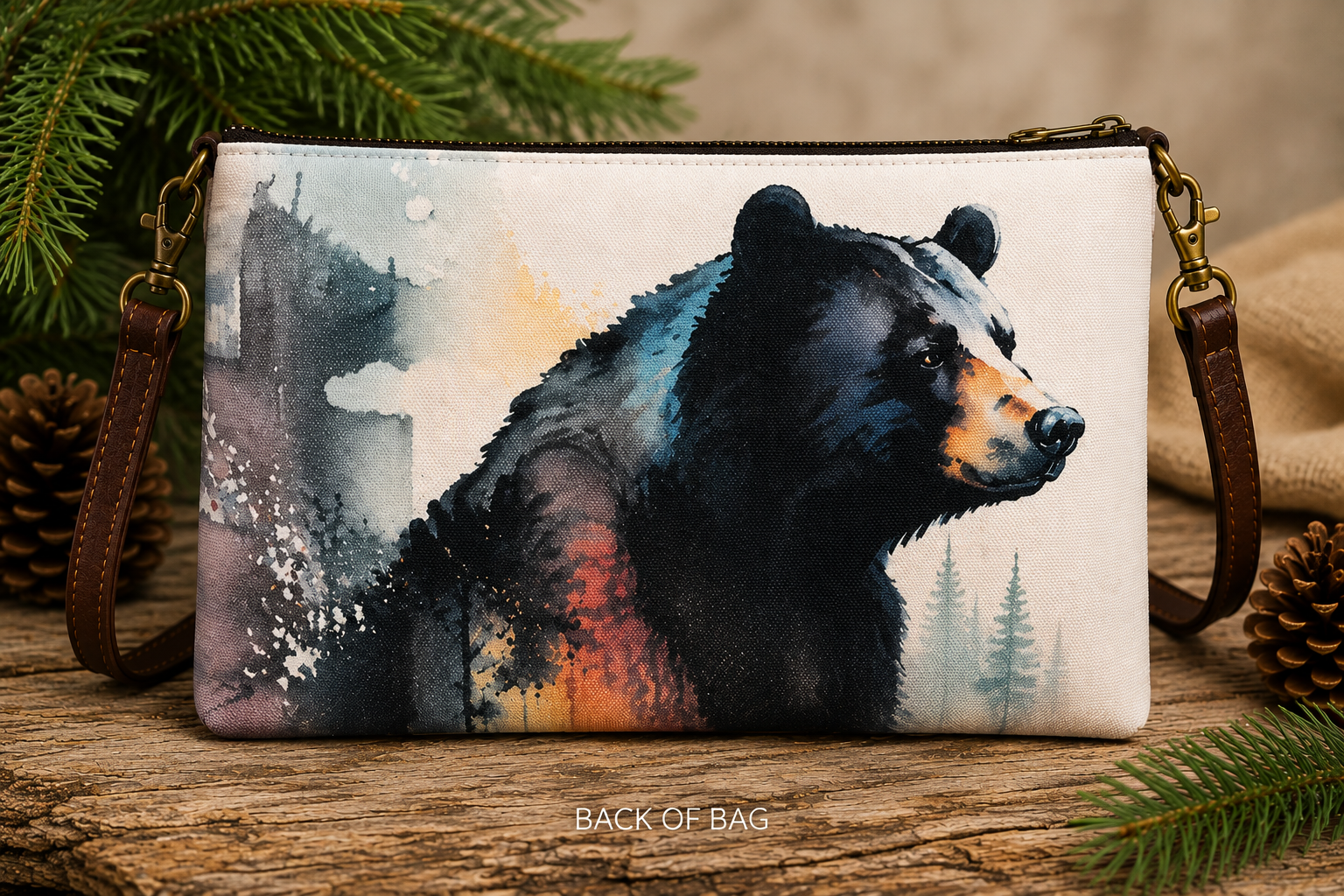 Back view of canvas crossbody bag with watercolor black bear and pine tree design, surrounded by pine branches and pinecones on a wood surface — Anngoti