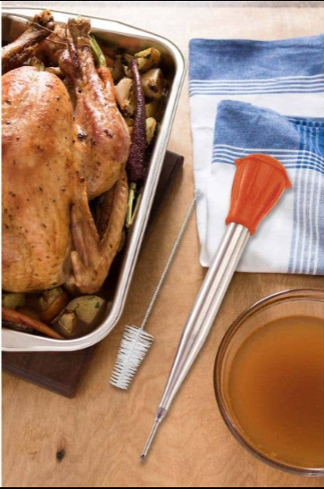 Overhead view of a whole roasted chicken in a roasting pan with the HIC baster and cleaning brush beside a bowl of pan drippings on a blue striped kitchen towel, showing the kit in a real cooking setting.