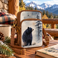 "Black bear in snow wildlife art canvas crossbody phone sling with leather-look straps and antique brass hardware, styled on a mountain cabin porch table with plaid blanket, pine branches, pinecones, and a camp mug against a snowy mountain backdrop"