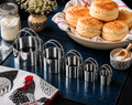 Set of 5 stainless steel biscuit cutters in graduated sizes displayed on a dark countertop beside a bowl of freshly baked biscuits, a Ball jar of flour, and a rooster kitchen towel — Fox Run