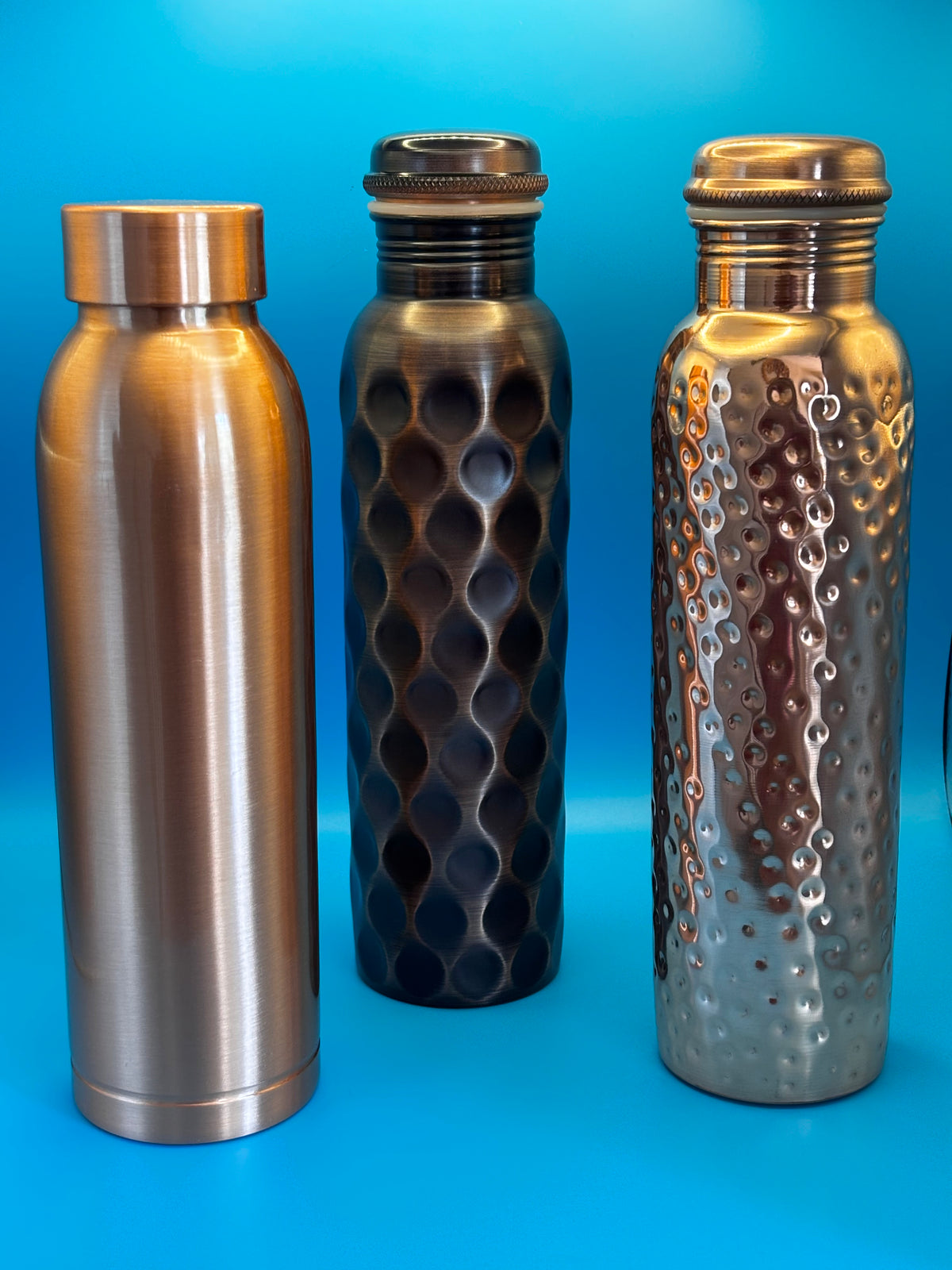 Perilla Home pure copper water bottle 1L — Cleo, Antique Diamond, and Hammered styles displayed together on a wooden shelf against a blue-gray background