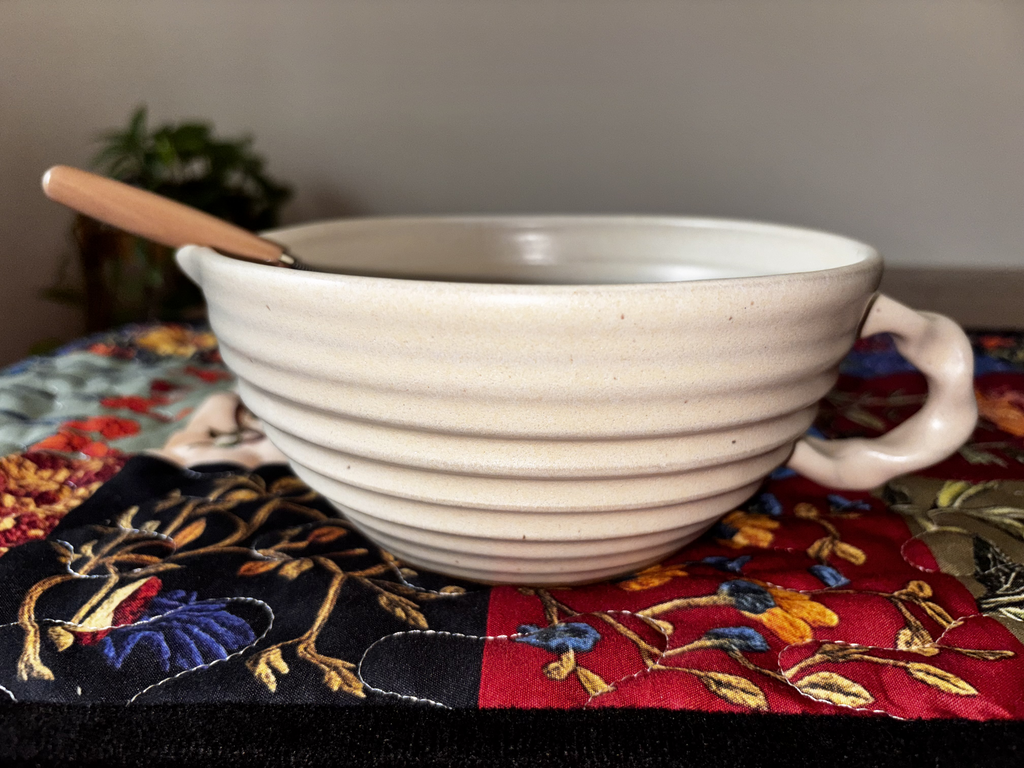  "Single Gravesco handmade cream ceramic mixing bowl on colorful quilted runner with whisk handle resting on left side"