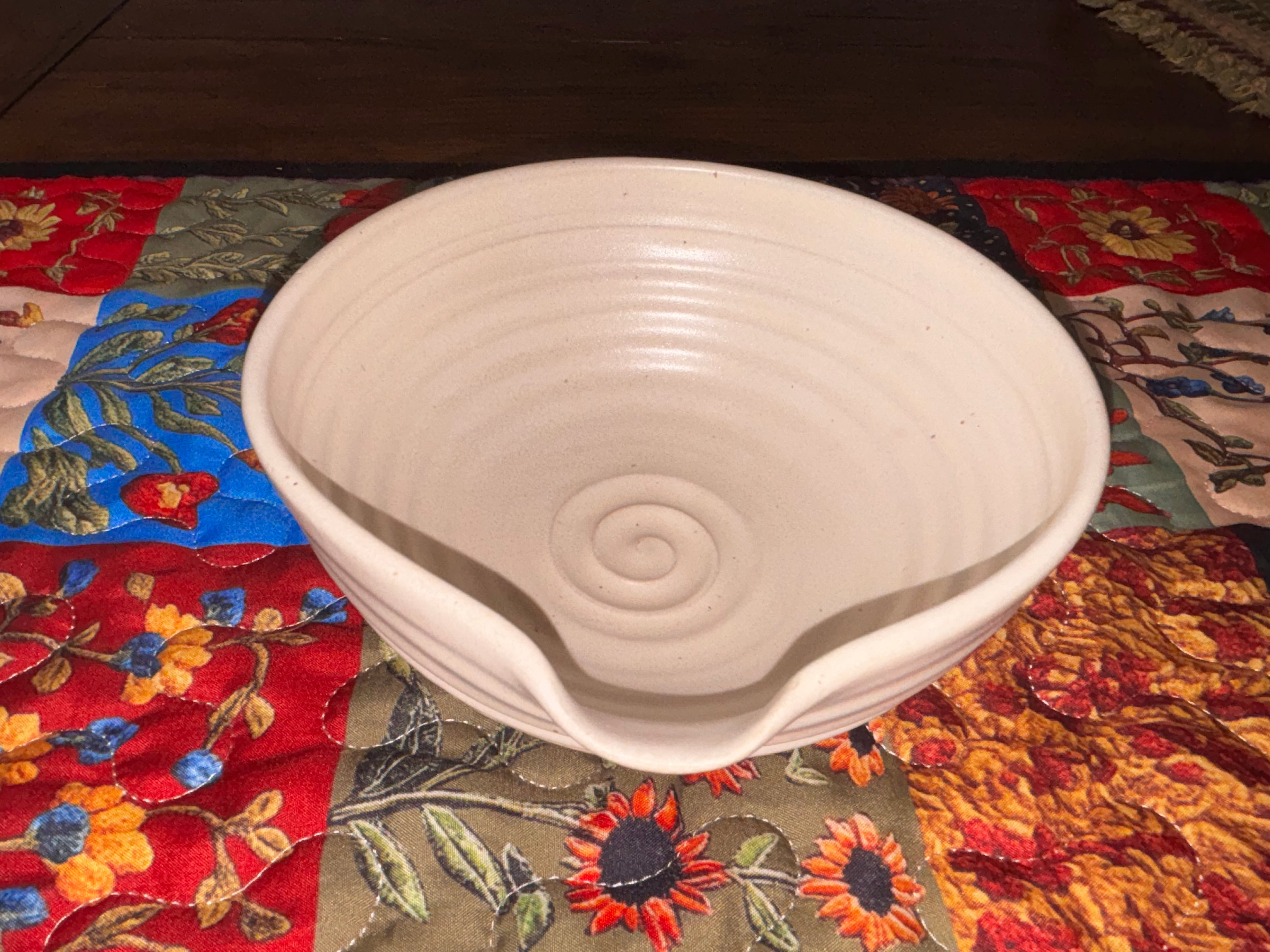 "Gravesco handmade cream ceramic mixing bowl on colorful quilted runner showing spiral interior and pour spout, angled view"