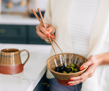 Lifestyle shot — woman holding bowl with chopsticks, mug in background: "Woman holding a handmade terracotta ceramic ramen bowl with chopsticks in a bright kitchen, with the coordinating Terra Cotta mug in the background"