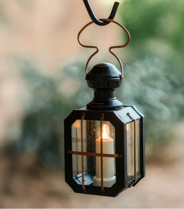 Barebones Box Candle Lantern hanging outdoors with lit candle glowing through seeded glass panels against soft green bokeh background"