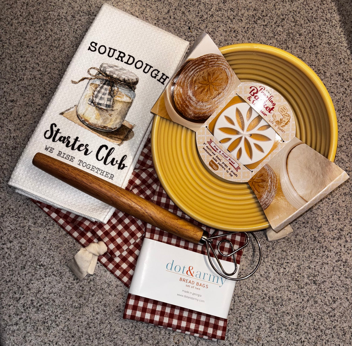 Bread-baking scene with dough in a yellow proofing basket, a checked bread bag draped over the edge, Starter Club towel and dough whisk.