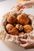 Hands lifting a large Bee's Wrap Bakery Lane beeswax wrap lined inside a bowl filled with assorted fresh-baked pastries including croissants, muffins, and a hot cross bun