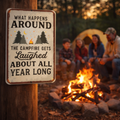 People sitting around a campfire with a sign that reads 'What happens around the campfire gets laughed about all year long' in a forest setting.
