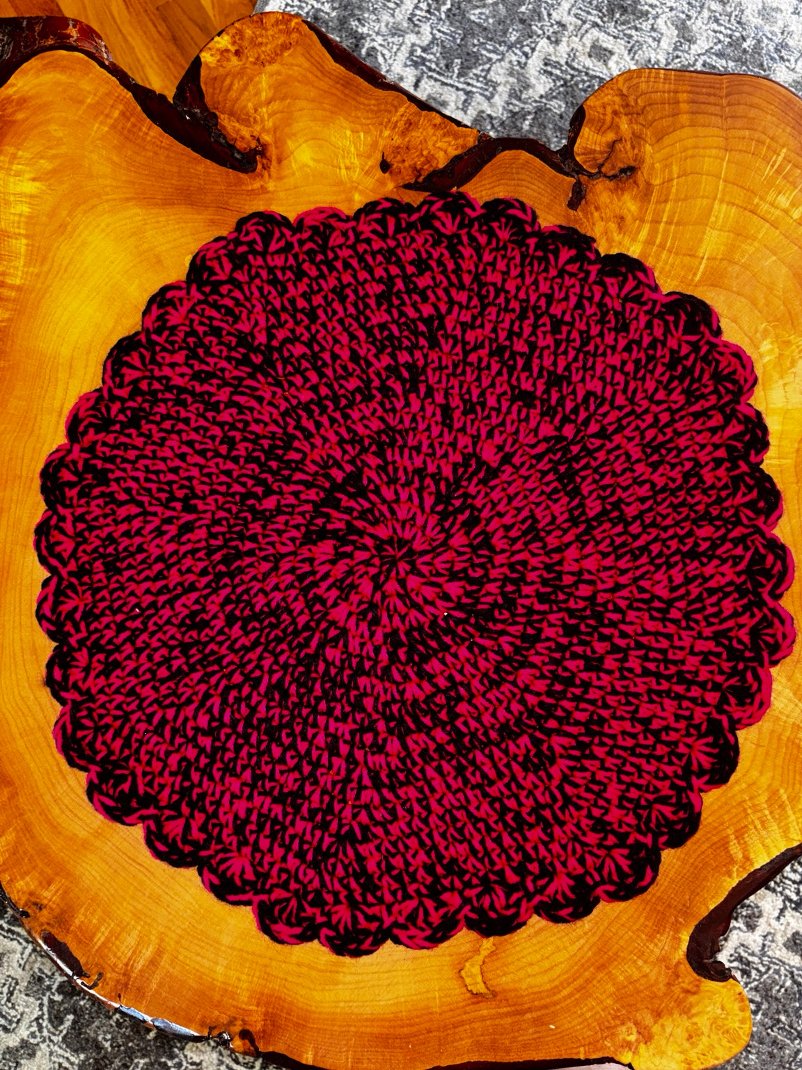 A round, hand-crocheted cat lounger in a deep magenta  and black pattern, displayed flat on a rustic live-edge wooden slice.