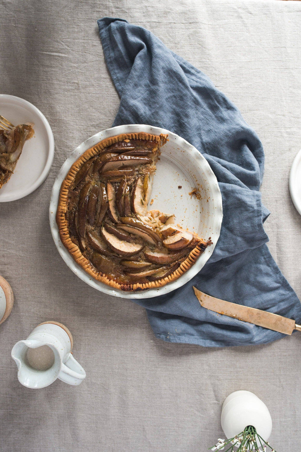 Overhead view of a partially sliced caramelized pear tart in a white speckled stoneware pie dish with ruffled edge, styled on a linen tablecloth with a blue napkin and knife