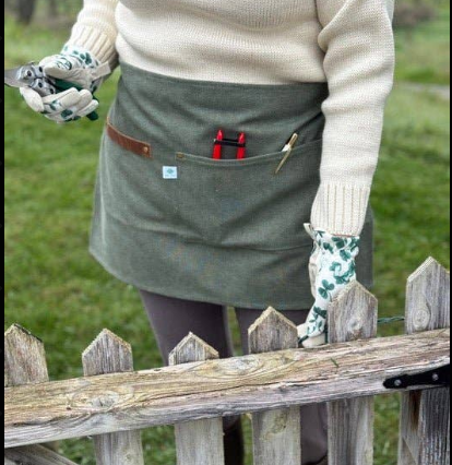 Celtic Farm hunter green waxed canvas half apron worn outdoors with pruners and tools in pocket, garden setting with white picket fence