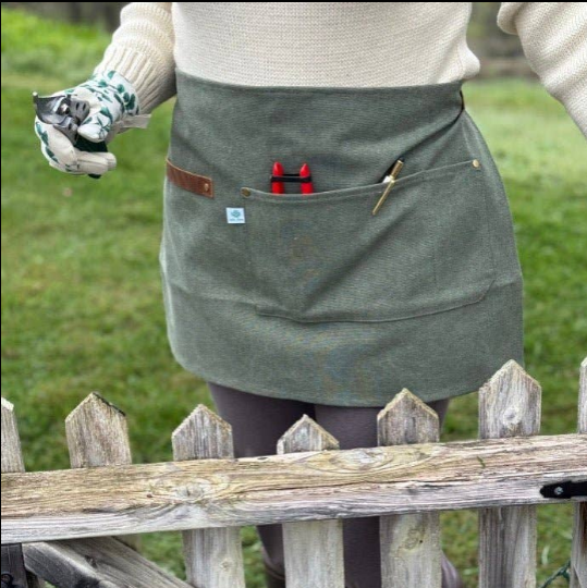 Celtic Farm hunter green waxed canvas half apron worn in garden with tools in pocket, gardening gloves in hand, wooden picket fence in background