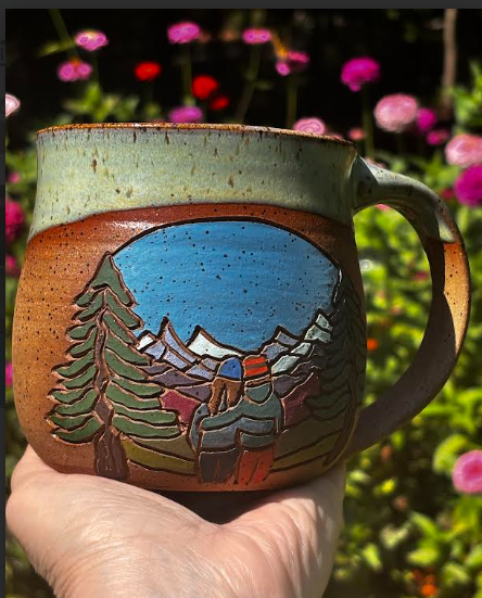 Forest Jeannie Pottery handmade stoneware mug held in hand showing hand-carved couple viewing mountain landscape with pine trees, with colorful garden flowers in background