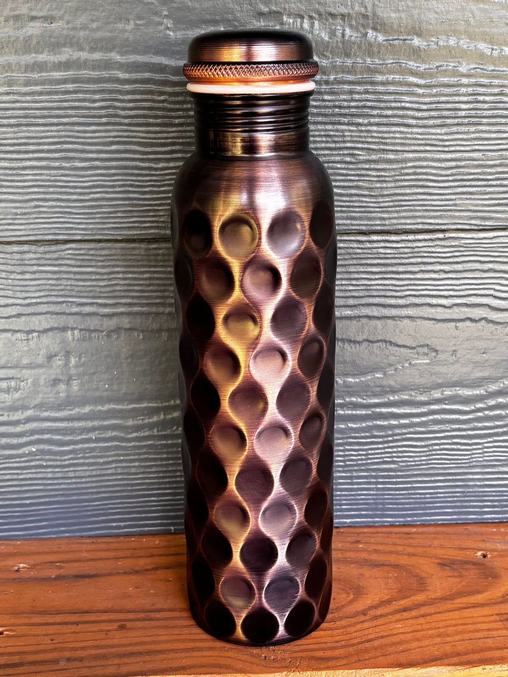 Perilla Home Antique Diamond pure copper water bottle 1L — deep dark copper with diamond-cut pattern and knurled cap, on a wooden surface against a blue-gray wood background