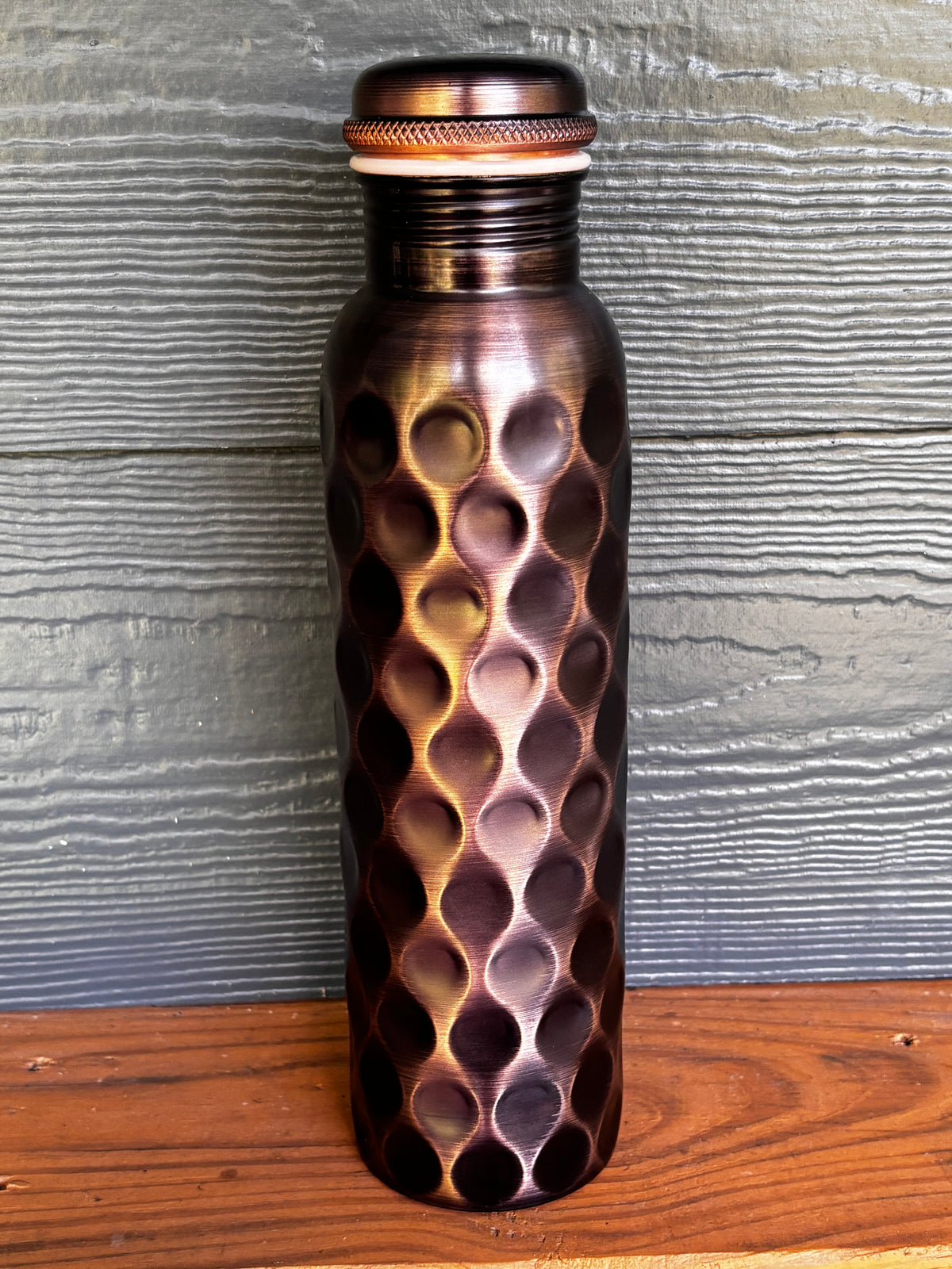 Perilla Home Antique Diamond pure copper water bottle 1L — deep dark copper with diamond-cut pattern and knurled cap, on a wooden surface against a blue-gray wood background