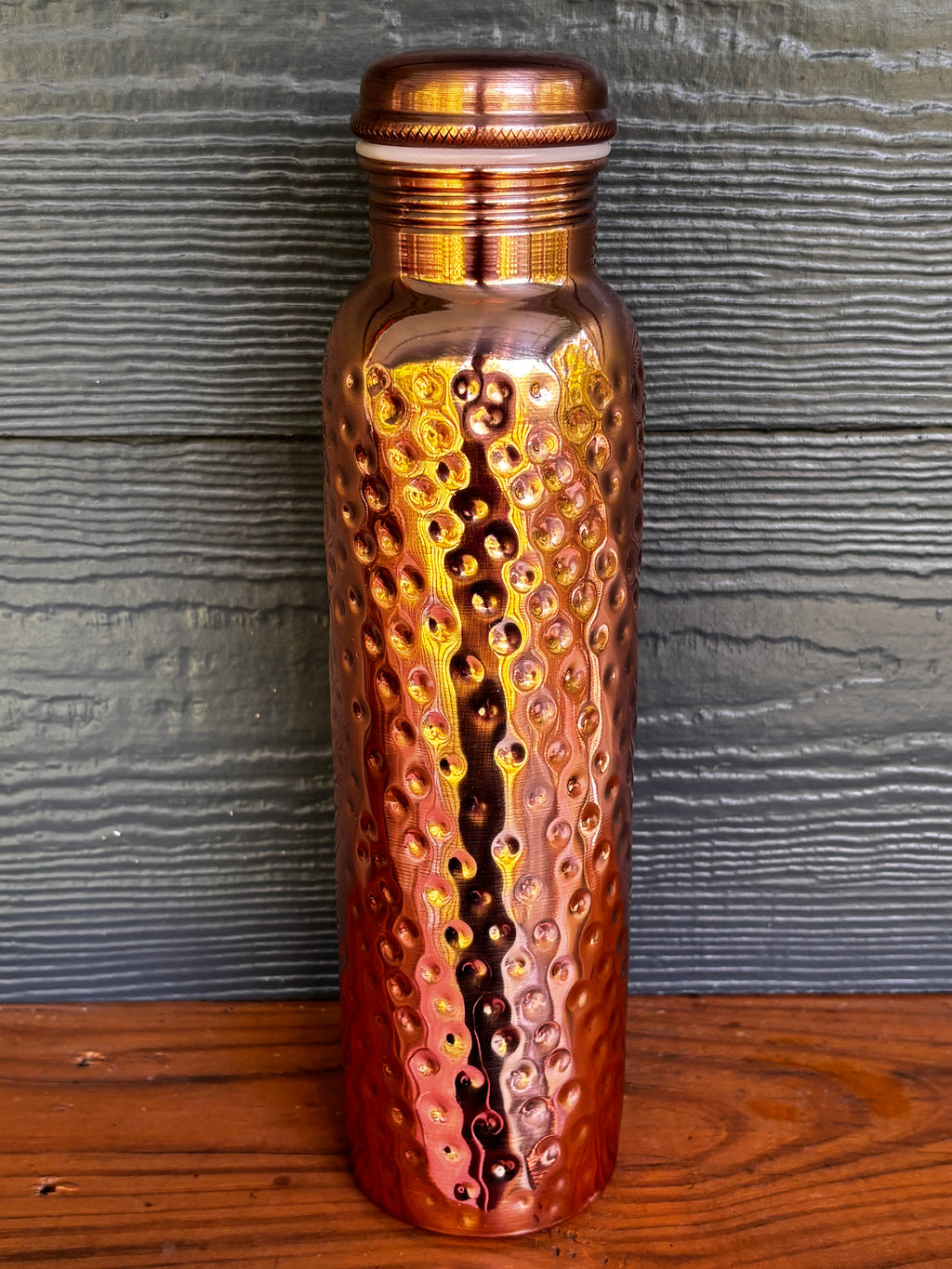 Perilla Home Hammered pure copper water bottle 1L — rich warm copper tones with hand-hammered texture and knurled cap, on a wooden surface against a blue-gray wood background