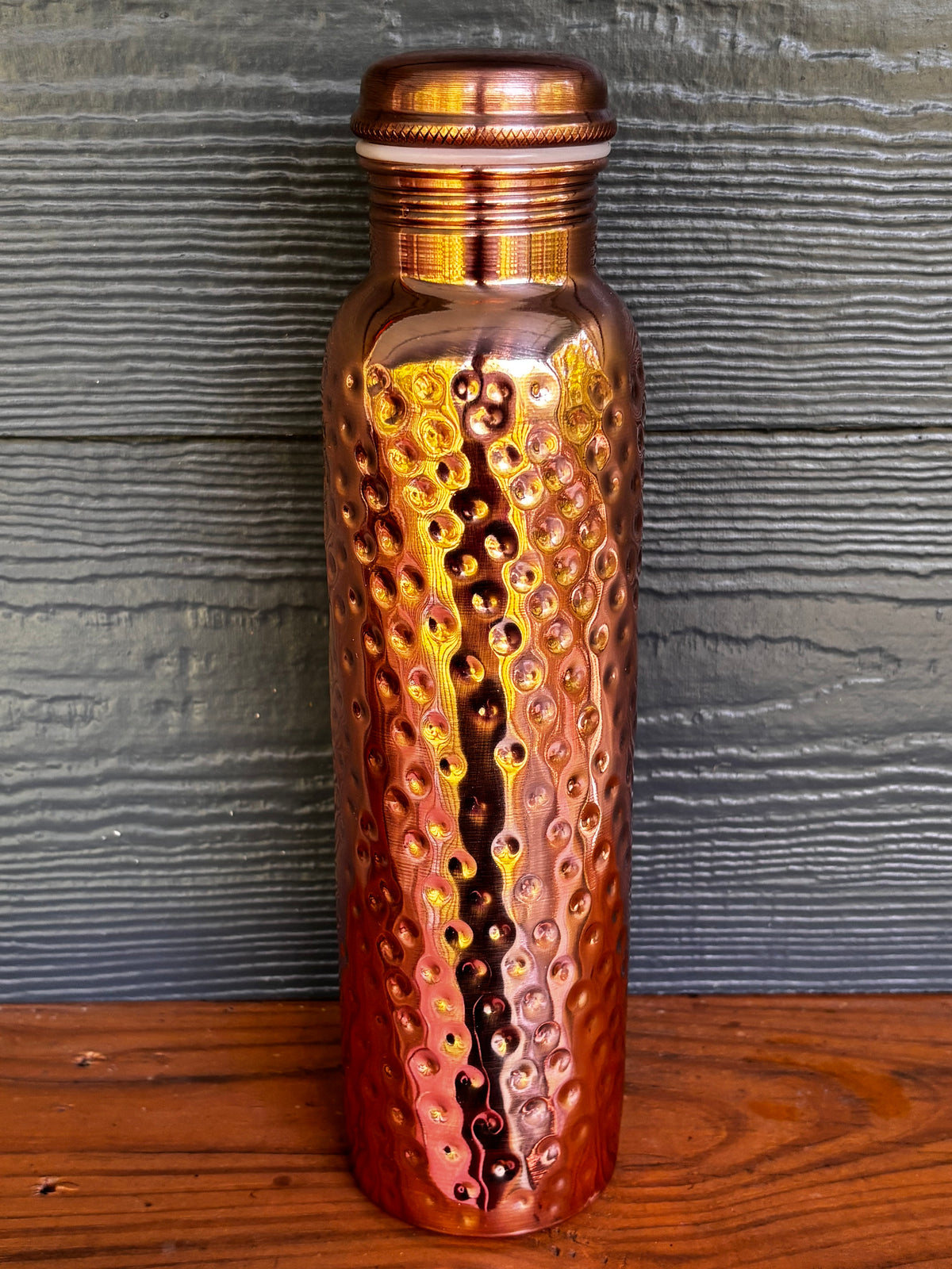 Perilla Home Hammered pure copper water bottle 1L — rich warm copper tones with hand-hammered texture and knurled cap, on a wooden surface against a blue-gray wood background