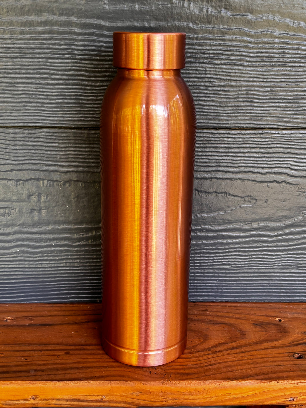 Perilla Home Cleo pure copper water bottle 1L — smooth, modern design in warm copper tones, standing on a wooden surface against a blue-gray wood background