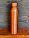 Perilla Home Cleo pure copper water bottle 1L — smooth, modern design in warm copper tones, standing on a wooden surface against a blue-gray wood background