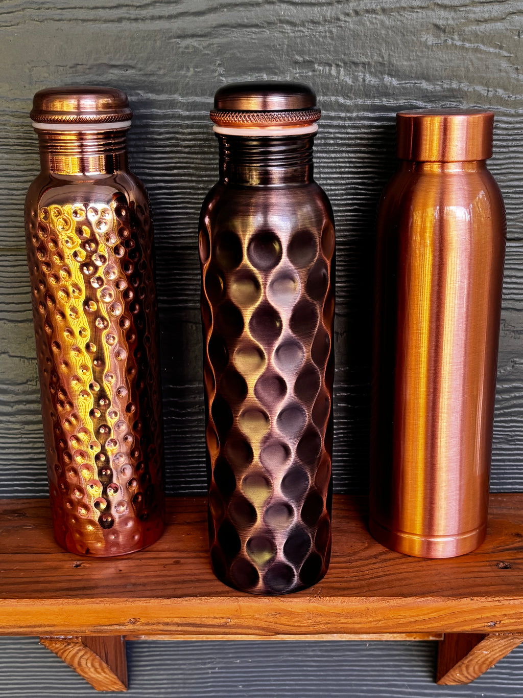 All three Perilla Home pure copper water bottles 1L — Hammered, Antique Diamond, and Cleo styles side by side on a wooden shelf against a dark blue-gray background