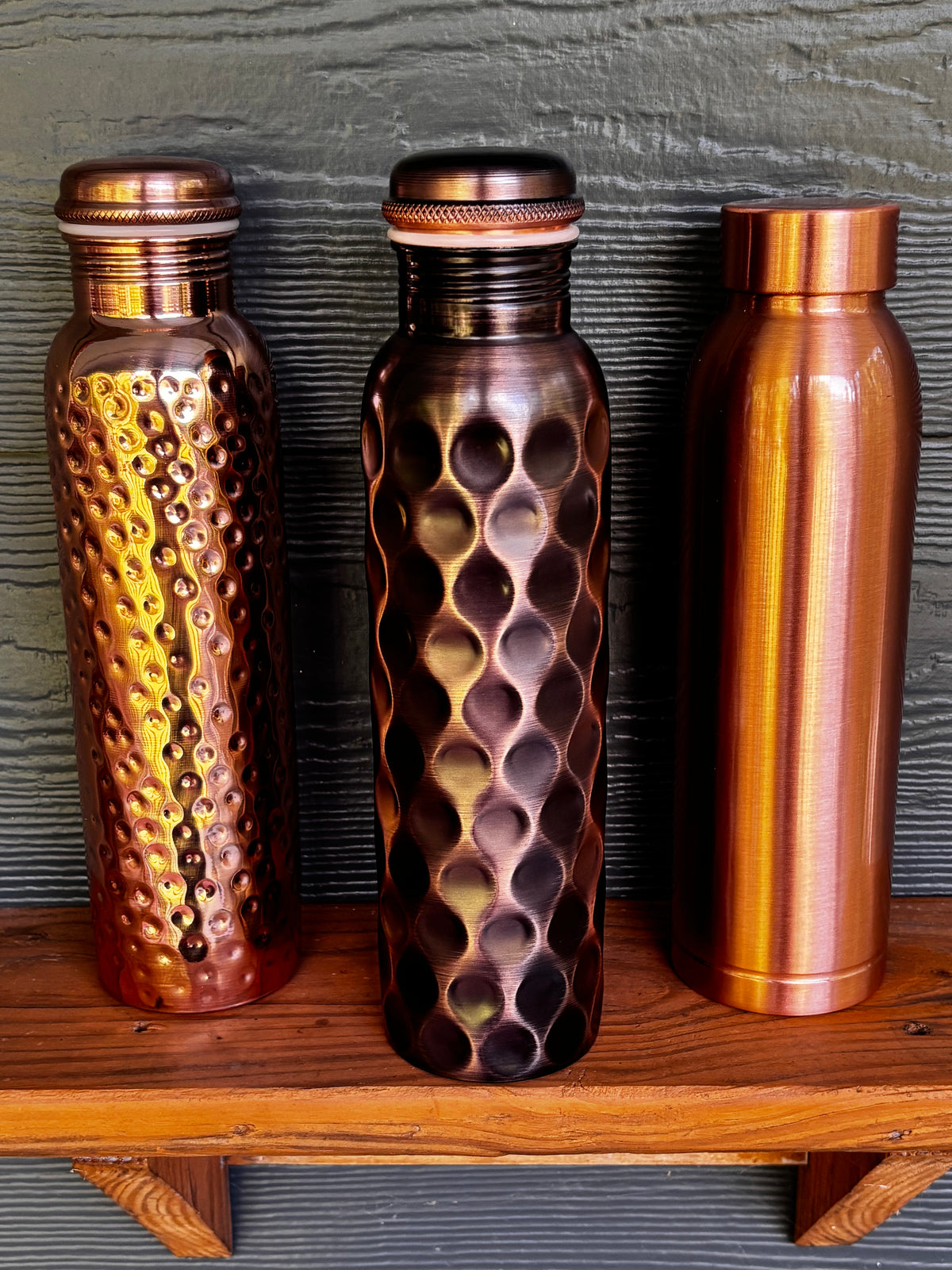 All three Perilla Home pure copper water bottles 1L — Hammered, Antique Diamond, and Cleo styles side by side on a wooden shelf against a dark blue-gray background