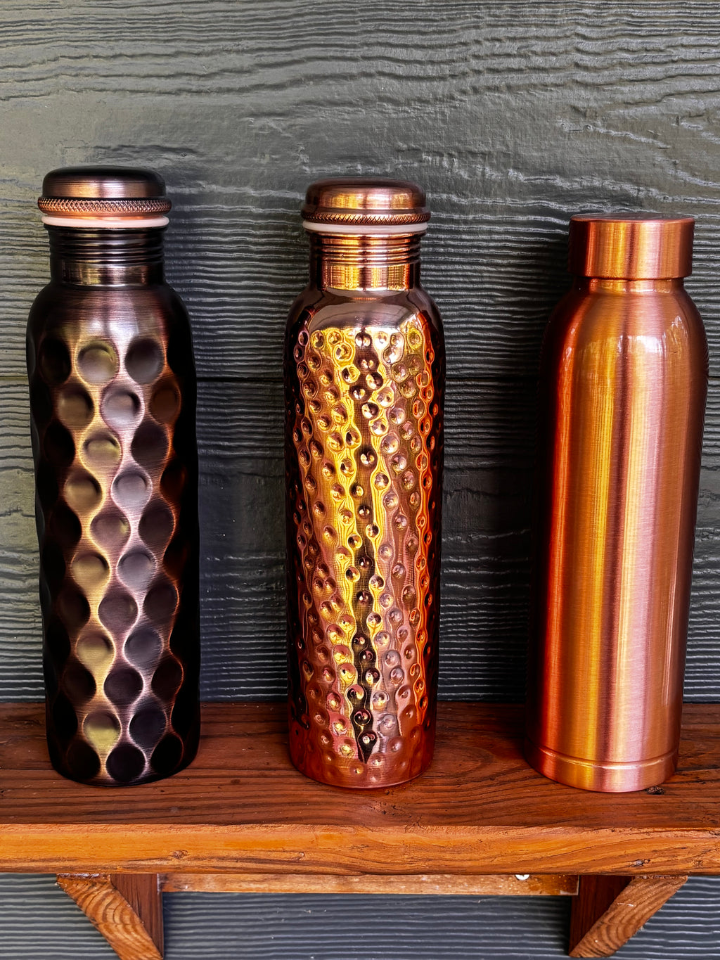 Perilla Home pure copper water bottle collection 1L — Antique Diamond, Hammered, and Cleo styles standing together on a wooden shelf, showing the range of artisan textures and copper tones