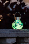 Allsop Tea Lantern teal solar lantern glowing green at night on a stone ledge with dark foliage silhouette in background"