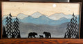 A rectangular wooden wall art piece featuring a painted mountain landscape with layered blue and gray peaks, a pale cream sky, black evergreen trees, and black bear silhouettes at the bottom. The piece is photographed against a dark background.