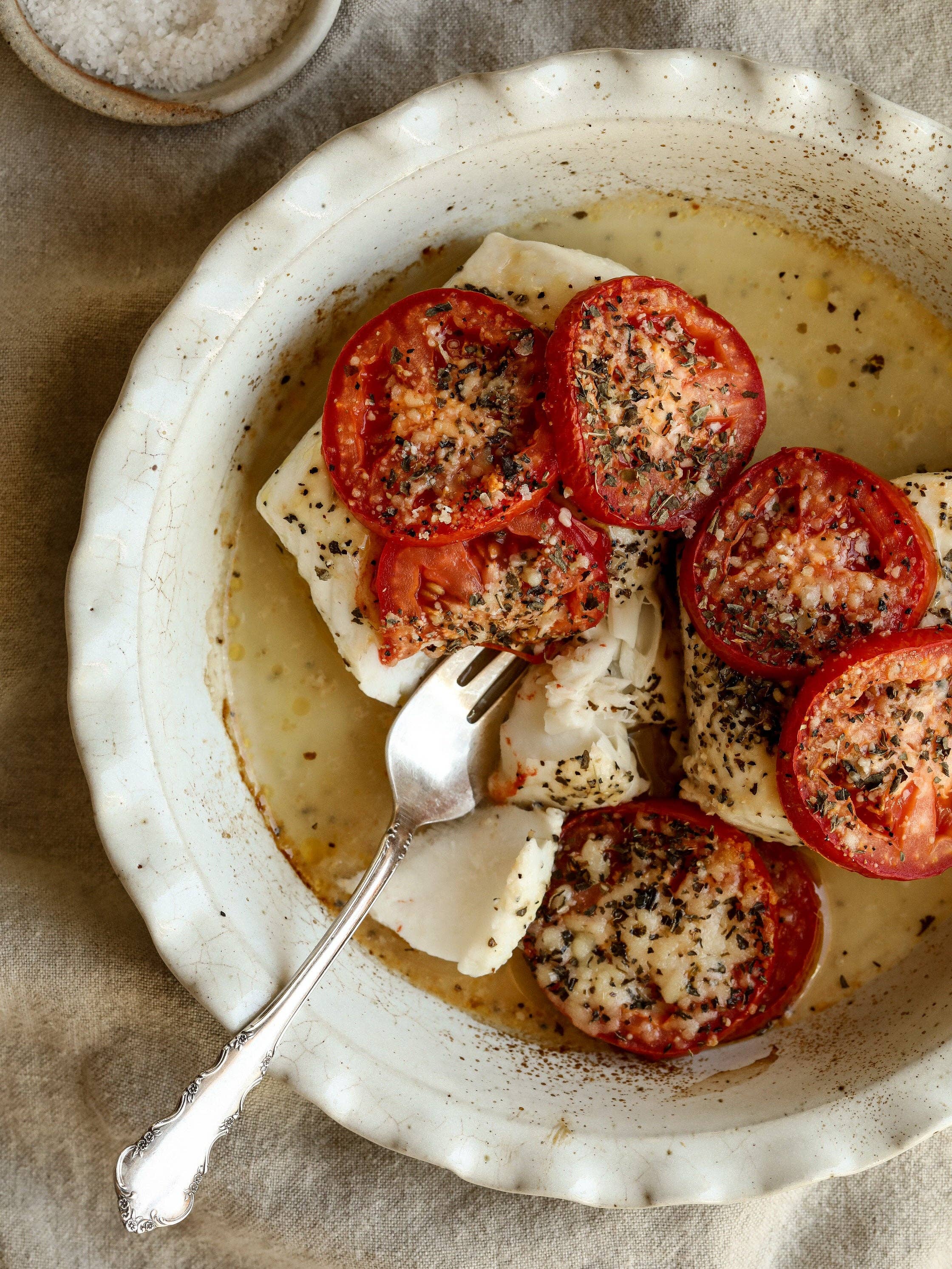 Overhead view of roasted tomatoes with herbs and mozzarella in a white speckled stoneware pie dish with ruffled scalloped edge, styled on a linen surface with a silver fork