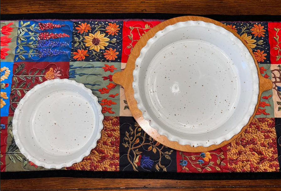 Two handmade speckled white stoneware pie dishes in small and large sizes displayed on a colorful floral quilted table runner"
