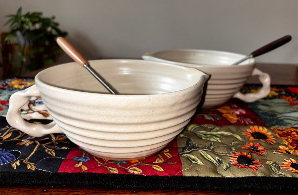 "Two Gravesco handmade wheel-thrown cream mixing bowls with whisks on a colorful floral quilted runner, side view"