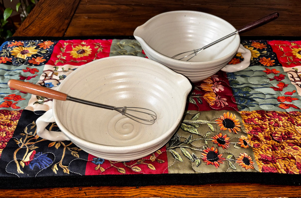 "Two handmade cream ceramic mixing bowls with pour spout and handle, styled with whisks on a colorful quilted table runner"