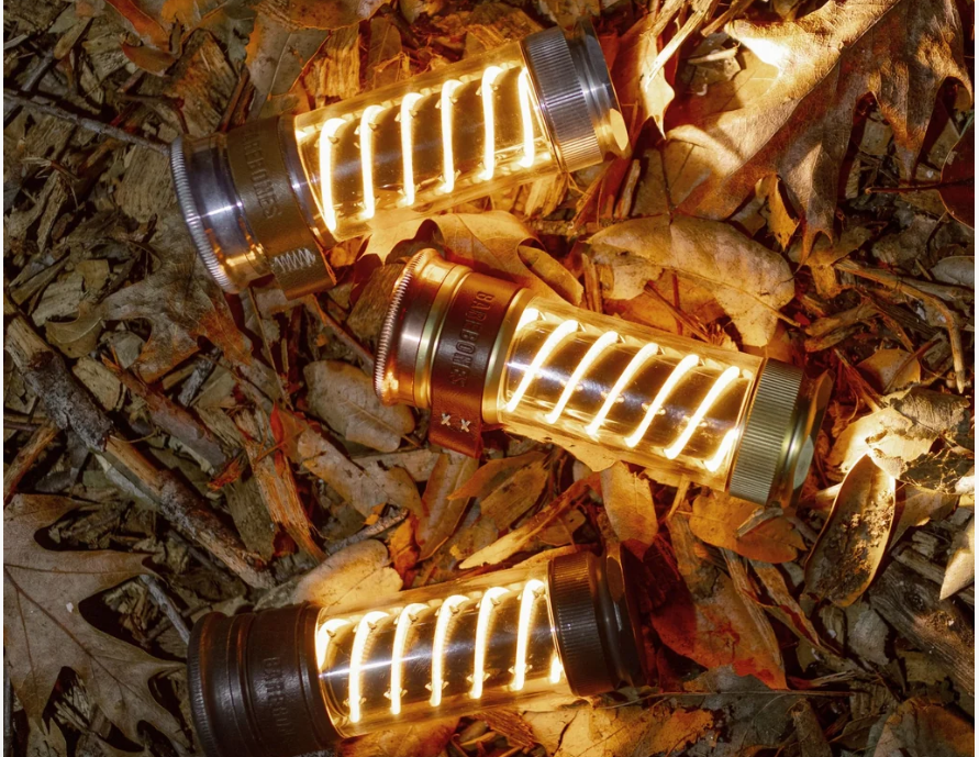 Three Barebones Edison Light Sticks in bronze and silver finishes glowing with warm amber light scattered across autumn leaves